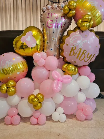 An elaborate balloon bouquet for a baby girl celebration. It includes various foil balloons like 'Hello World' and 'It's a Girl' mixed with pink, white, and gold latex balloons.