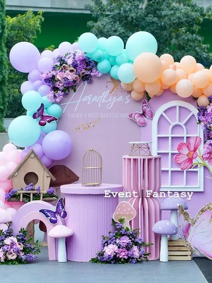 A beautiful pastel-colored setup for Aaradhya's butterfly party. This photo corner features a large pink backdrop, a balloon arch in shades of purple, teal, and peach, and whimsical props like a birdhouse and mushrooms.