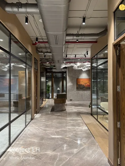 The main corridor of 'Project LH,' a 4,000 sq ft corporate office in Worli. The design uses a combination of marble flooring, glass partitions, and exposed ducting to create a sleek, modern, and dynamic pathway through the workspace.