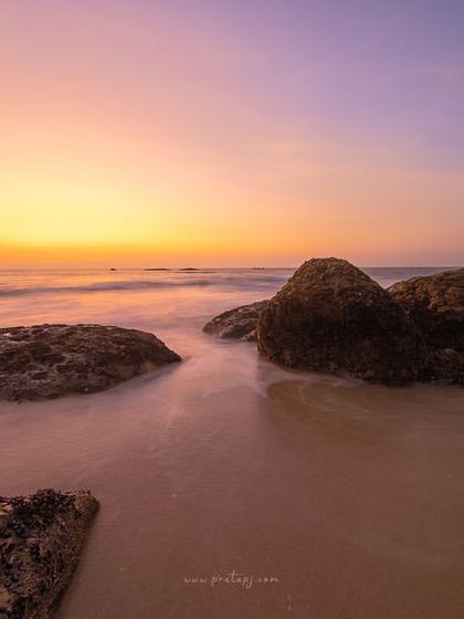 A long exposure of the sea at Padubidri beach during a colorful sunset. The slow shutter speed turns the waves into a smooth, silky surface, highlighting the stillness of the rocks.