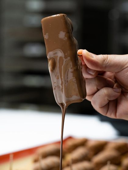 An action shot from Chef Karim Bourgi's masterclass, capturing the satisfying drip of chocolate from a freshly coated bar.