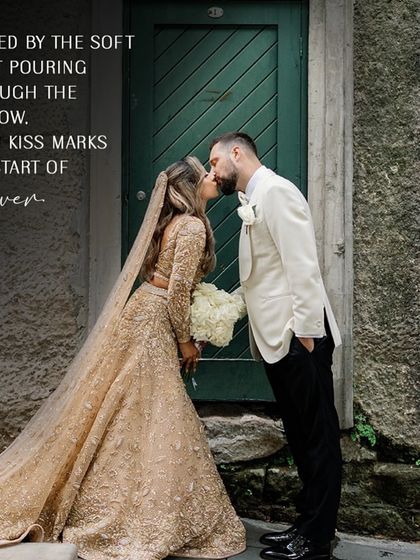 Framed by the soft light pouring through the window, their kiss marks the start of forever. A beautiful shot showcasing the bride's embellished golden lehenga and the groom's sharp tuxedo.