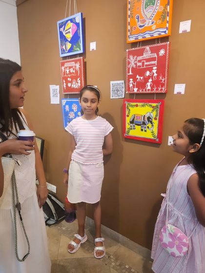 A young artist stands proudly next to her work, sharing her creative journey with an attentive visitor at the exhibition.