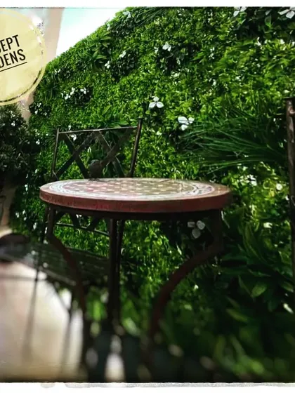 A simple cafe-style seating arrangement set against a backdrop of our artificial green wall with small white flowers. This creates a charming and maintenance-free outdoor cafe feel.