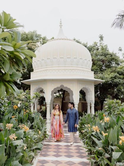 Shikha and Rupen walking hand-in-hand through a garden path at a palace, a serene and romantic moment.