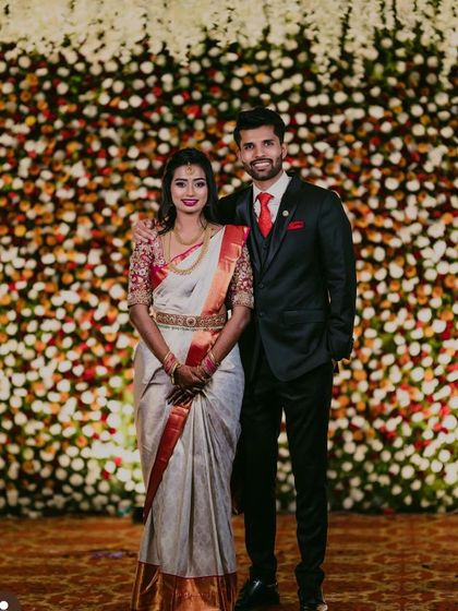 A happy couple posing in front of a dense floral wall. The mix of white, pink, and orange flowers creates a warm and inviting atmosphere for the reception.