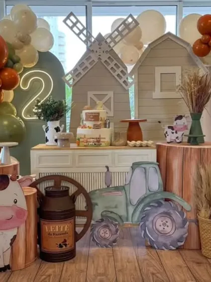 A charming farm animal theme for a second birthday. The setup includes a barn backdrop, a windmill, a tractor prop, and cute cow and horse cutouts.