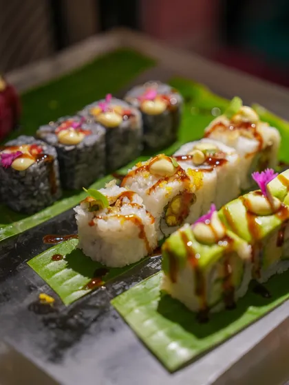 A variety of sushi rolls, including avocado-topped and classic maki, drizzled with sauces and garnished with edible flowers.