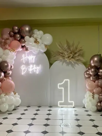 A minimalist and elegant first birthday setup with white backdrops, a neon sign, a "1" marquee number, and a balloon arch in muted pink, brown, and white.