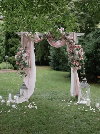 A romantic garden arch with soft pink drapes and floral arrangements, flanked by lanterns for a dreamy, rustic feel.