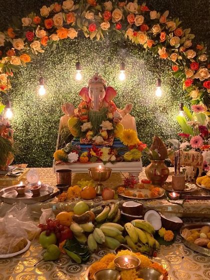 A rustic and earthy Ganpati decoration with a green foliage backdrop and an arch of orange and white flowers. The exposed Edison bulbs add a warm, vintage touch to the festive setup.