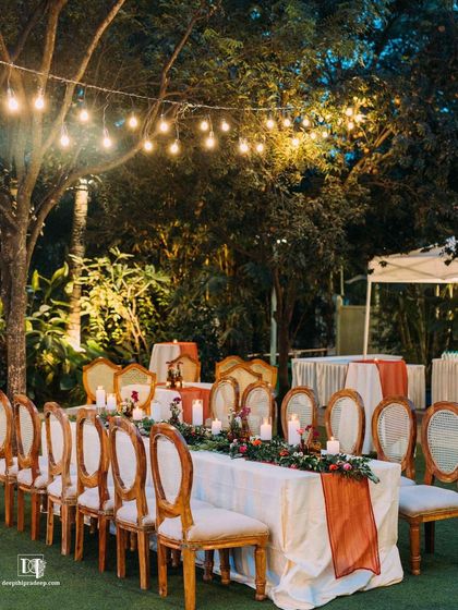 A beautiful long-table setup for an evening dinner. The string lights overhead and the lush trees surrounding the lawn create a magical dining experience.