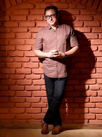 A full-length portrait of Sanjev Sharma against a brick wall. The simple background and strong pose create a confident and grounded image.