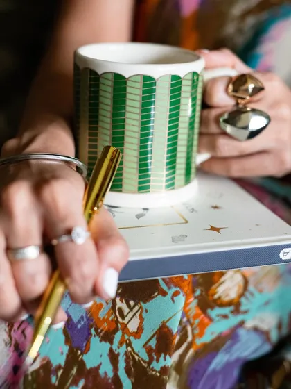 A detailed close-up of products in use for Patrika Gift Shop. This shot focuses on the hands holding a pen and a beautifully designed mug, highlighting the tactile experience of the brand's items.