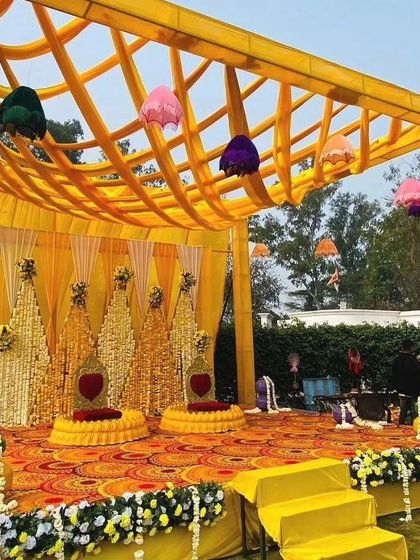 A full view of the grand Haldi mandap setup. The yellow canopy with hanging umbrellas complements the floral stage, creating a cohesive and impressive look for a large-scale outdoor celebration.