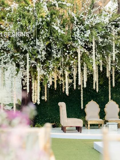 A detailed view of the mandap stage, framed by a cascade of white flowers hanging from the trees. The simple seating arrangement maintains a focus on the natural beauty of the surroundings.