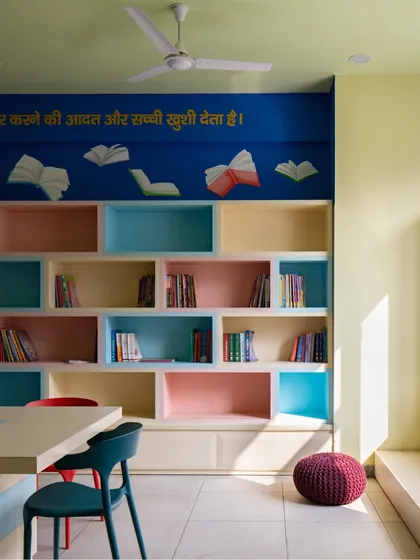 A cozy library corner with colorful, recessed bookshelves and a reading nook bathed in sunlight. We use color and light to create warm, uplifting spaces that make people feel at ease.