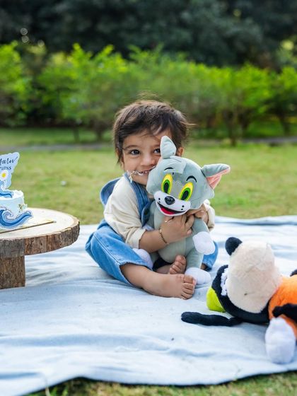 Little Aariv, with cake on his face, happily hugs his Tom and Jerry plushie during his first birthday celebration. A picture of pure, simple childhood joy.