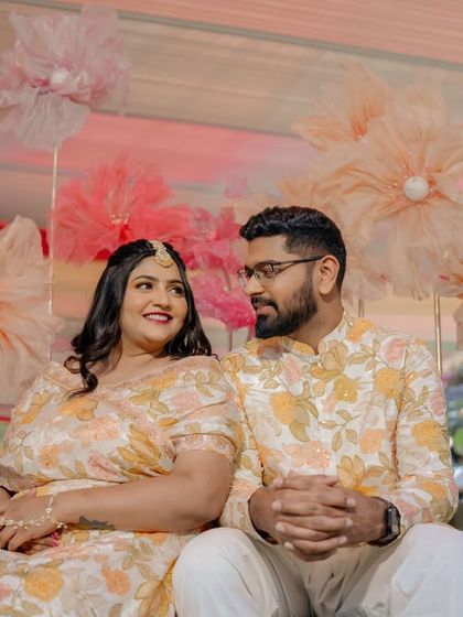A sweet moment between the couple during their Mehandi ceremony. The backdrop of large, handmade paper flowers in pastel shades adds a touch of whimsy and romance to the setting.