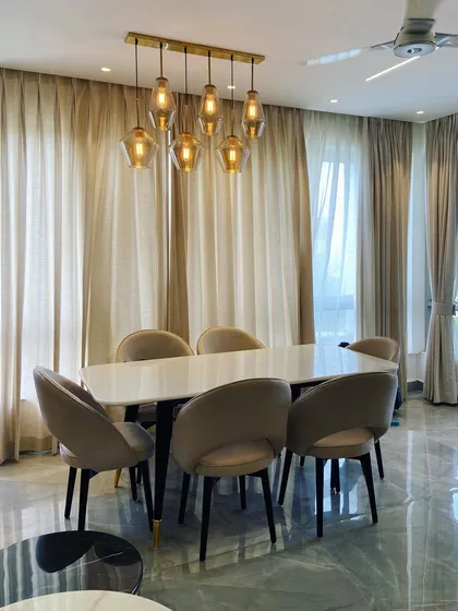 This view captures the dining setup at the Noida residence, showing the relationship between the statement pendant lighting and the six-seater table. The neutral, earthy tones of the chairs and curtains create a calm and cohesive look.