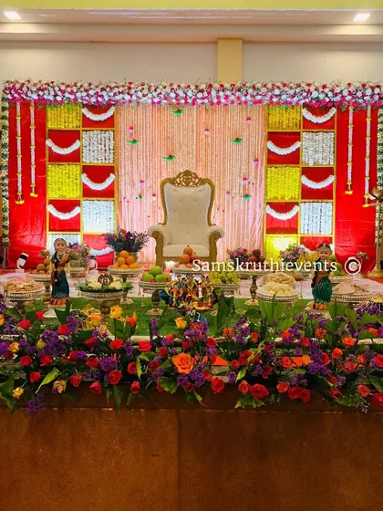 A bright and colorful Seemantha decoration with a red and yellow theme. The stage is filled with a variety of flowers and a neat arrangement of offerings, creating a joyful and traditional atmosphere.
