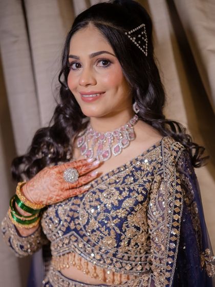 A modern and chic sangeet look. The bride is wearing a deep blue embroidered lehenga, and her look is completed with soft waves, a trendy triangular hair clip, and a sparkling diamond necklace.