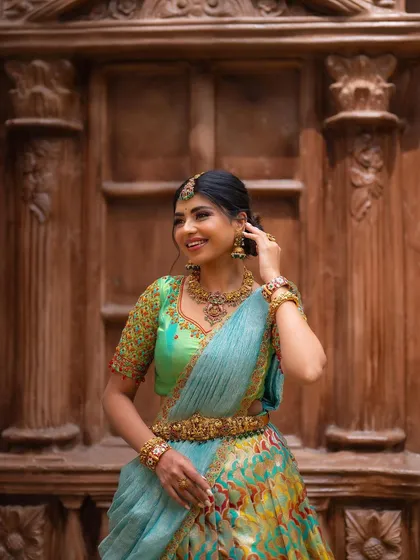 A candid, smiling portrait of a model in a designer half saree. The intricate wooden background adds texture and depth to this beautiful traditional shot.