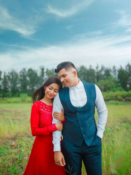A serene and romantic portrait set in a lush green field under a beautiful blue sky. The couple's formal attire creates a wonderful contrast with the natural surroundings, resulting in a very elegant pre-wedding photograph.