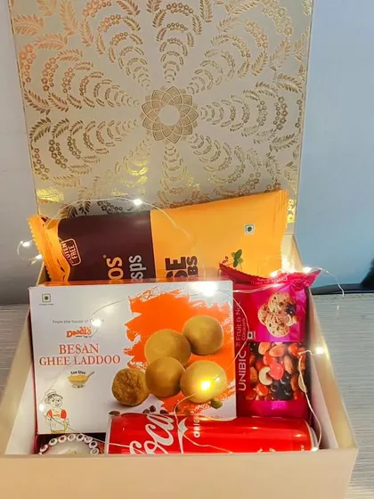 A corporate Diwali hamper featuring Besan Ghee Laddoos, cheese crisps, a can of Coke, and a trail mix, lit with fairy lights.