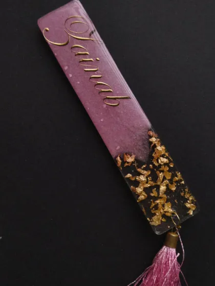 A close up of a purple and gold flake bookmark for 'Zainab', with a matching pink tassel.