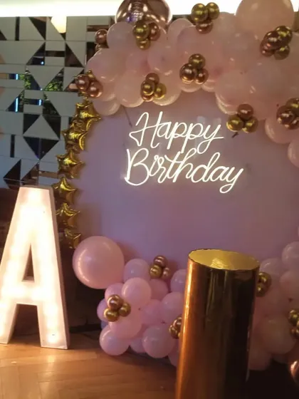 A simple and elegant backdrop in a restaurant setting. The pink balloon arrangement is paired with a 'Happy Birthday' neon sign and a large light-up initial 'A'.