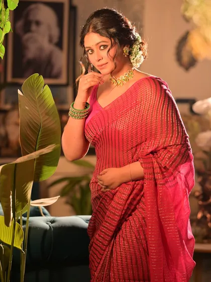 A festive portrait for 'Shubho Noboborsho' (Bengali New Year). The model is dressed in a shimmering pink saree, with her hair adorned with flowers, set in a warmly lit, decorated room.