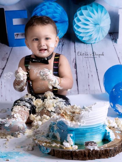 The delightful aftermath of a cakesmash, with the baby boy covered in blue and white frosting, thoroughly enjoying the demolition of his cake.