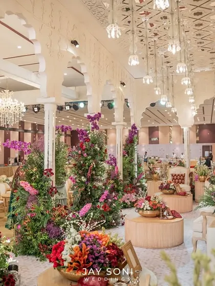 A breathtaking view of the lounge area at the "Garden of Vows" wedding, filled with towering floral arrangements and elegant seating under a ceiling of hanging lights and traditional arches.