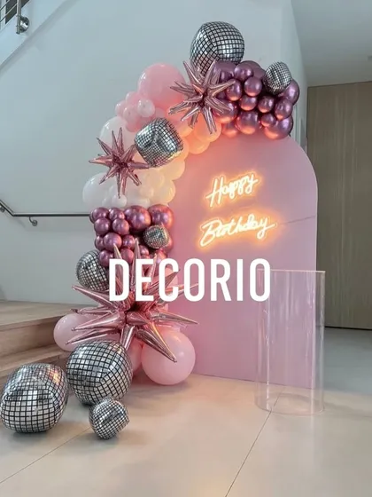 A trendy and fun birthday setup with a pink arched backdrop and neon sign. The balloon arrangement includes pink, white, and metallic balloons, accented with disco balls and starburst foils for a party vibe.