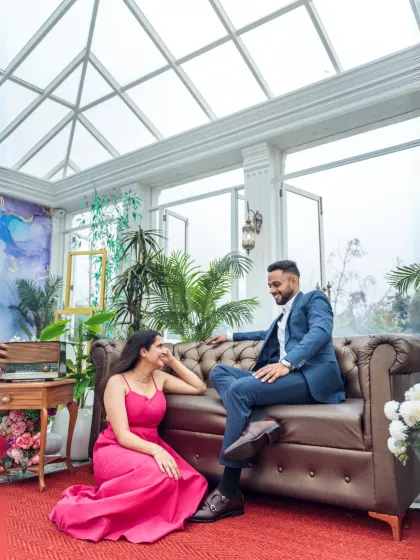 A relaxed and happy moment inside a sun-filled glasshouse. The couple shares a laugh on a leather chesterfield sofa, surrounded by lush greenery, creating a fresh and modern feel.