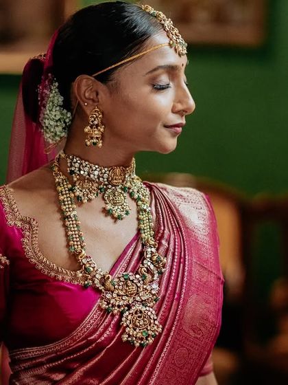 A profile shot of a bride, her intricate jewelry and the rich color of her saree highlighted against the deep green wall of a suite.