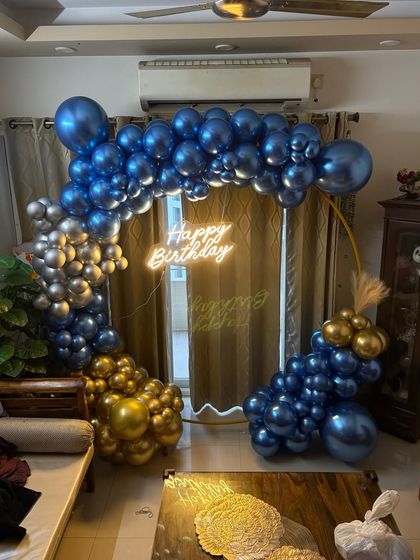 This is a great example of how we can create an elegant party atmosphere right in your living room. The setup includes a golden ring arch decorated with blue and gold chrome balloons and a 'Happy Birthday' neon sign.