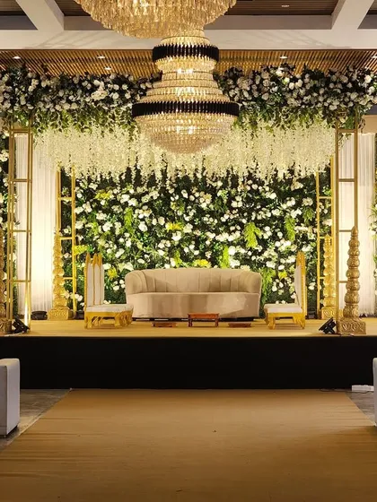 An elegant and lush reception stage featuring a floor-to-ceiling wall of fresh greenery and white florals. The design is elevated by cascading wisteria from above and magnificent crystal chandeliers, creating a look of timeless sophistication.