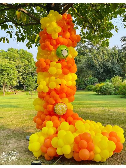 A tree on the lawn is decorated with a cascade of orange and yellow balloons, with kiwi-themed balloon details, tying the natural surroundings into the fruity party theme.
