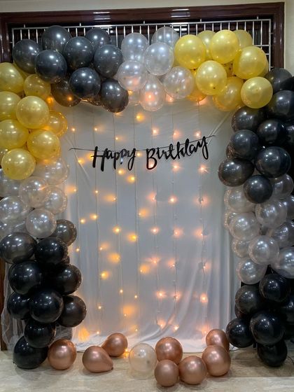 A classic birthday backdrop featuring a full balloon arch in gold, silver, and black. The 'Happy Birthday' banner is hung against a white curtain illuminated by a cascade of fairy lights.