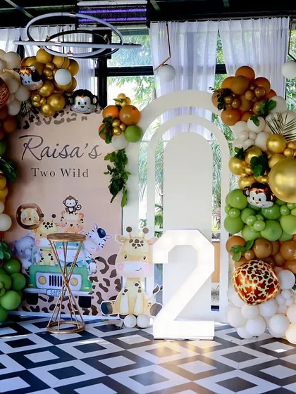 For this "Two Wild" jungle safari party, I created a fun and friendly backdrop with cute animal illustrations and a large number '2' marquee light. The scene is framed by organic balloon arches in earthy green and gold tones.