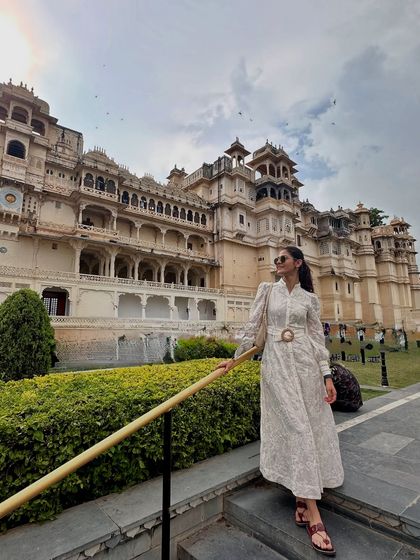 Standing before the City Palace in Udaipur in a white full-sleeve maxi dress. The classic silhouette and subtle print of the dress complement the grandeur of the palace architecture, creating a timeless and elegant vacation photo.