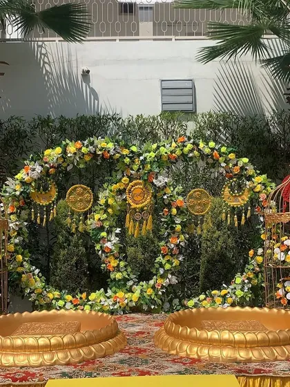 A detailed look at a Haldi seating area. The backdrop is a circular wreath of marigolds and greenery, complemented by golden birdcages and traditional 'urli' bowls for a complete look.