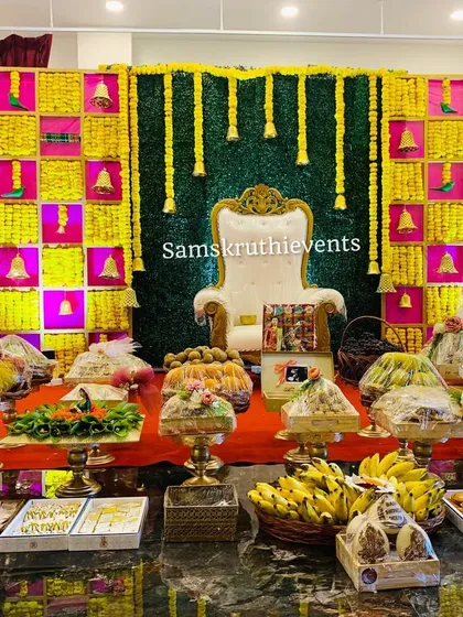 A vibrant baby shower backdrop in yellow and pink, with hanging marigold strings and traditional bells. The foreground shows a lavish display of covered offering trays, ready for the rituals.