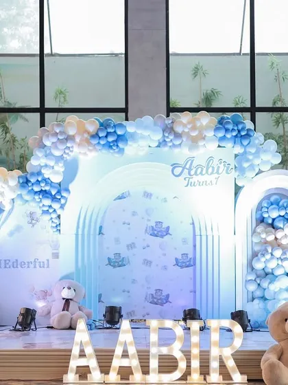This is a modern and dreamy teddy bear themed first birthday stage. The setup features a soft blue and white color palette, multiple balloon arches, illuminated marquee letters, and adorable teddy bears to create a cozy and celebratory scene.