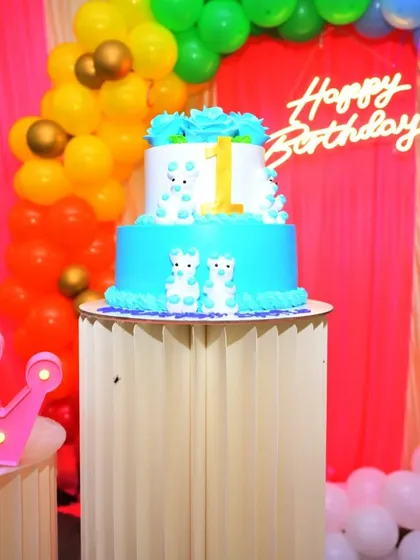A cute two-tiered cake for a first birthday. The cake is placed on a decorative pedestal that is part of the overall party setup.