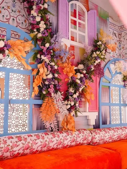 A vibrant seating nook at the Jaipur Haldi, with a bright orange sofa and floral-printed cushions. The backdrop features colorful window-like panels adorned with dense, multi-hued floral arrangements.