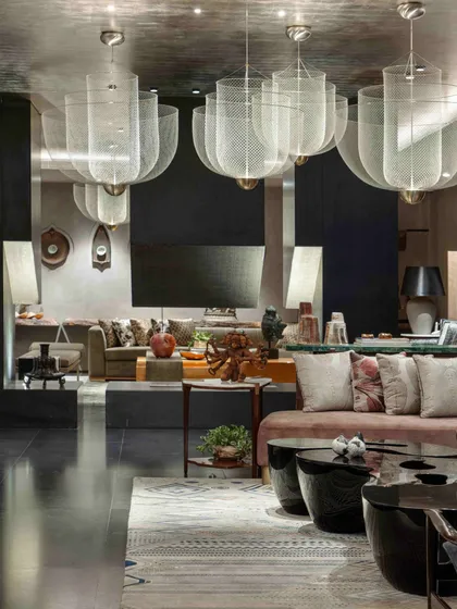 The formal living room in the basement of the Bammi Residence. The space is designed around a central fireplace and features large, dramatic mesh chandeliers, a dusty pink sofa, and a collection of art and artifacts.