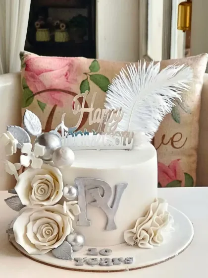 A sophisticated silver and white anniversary cake with sugar roses, a large feather, and monogrammed initials celebrating 20 years.